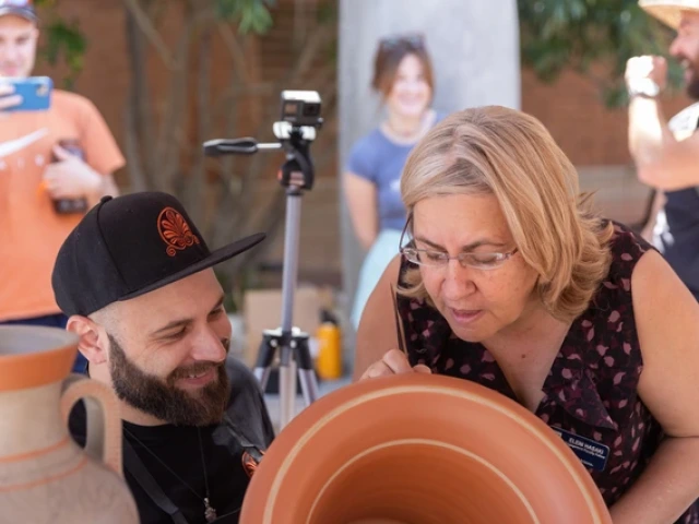 Eleni Hasaki looking over the shoulder of Roberto Paolini, an artist, to look at a reddish-colored vase that Paolini is painting a design on
