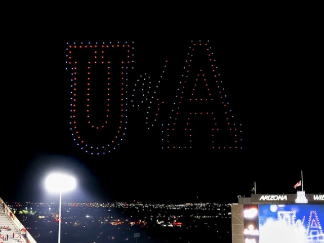 A nighttime drone show above Arizona Stadium forms a red, white, and blue "U of A" in the sky during a University of Arizona football game. The illuminated letters appear over the lit stands and scoreboard as the city lights of Tucson glow in the distance.