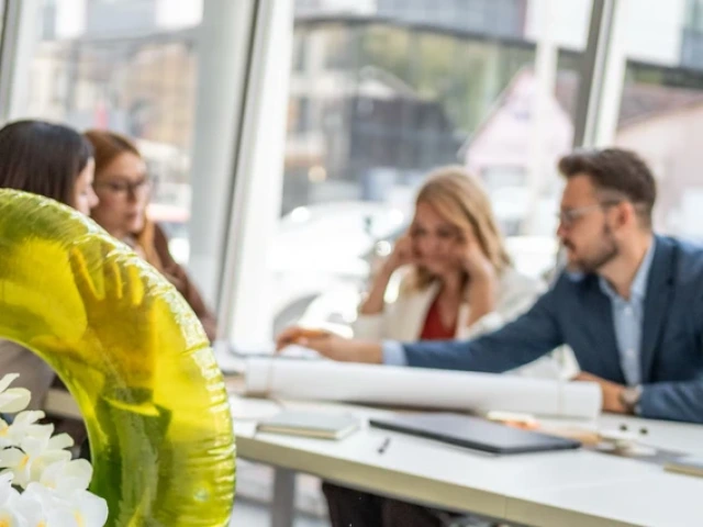 A man wearing a flower lei and a large yellow pool float laughs while sitting at a conference table, as colleagues in business attire hold a meeting in the background. The playful scene contrasts the relaxed mood of the man with the focused group discussion in a bright, modern office.