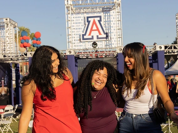 Three women smile and laugh in front of the Unviersity of Arizona logo.