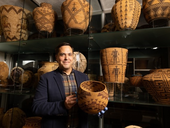 Edward A. Jolie standing while holding a handwoven basket in front of a wall with shelves of similar baskets