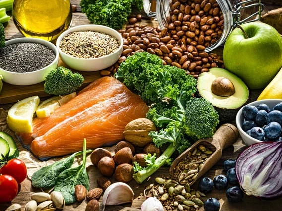 Colorful spread of healthy whole foods including a raw salmon fillet, avocado, blueberries, apple, banana, kale, broccoli, cherry tomatoes, red onion, lemon, chia seeds, quinoa, beans, assorted nuts and seeds, olive oil, and various leafy greens arranged on a rustic wooden surface. A glass jar spills over with pinto beans in the background, adding texture and warmth to the nutrient-rich display.