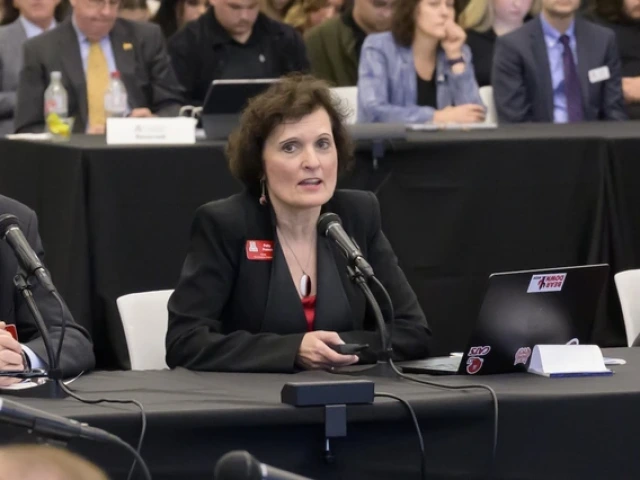 A woman in a black blazer speaks into a microphone while seated at a table during a formal meeting. She holds a small device in one hand, with an open laptop displaying several stickers in front of her. People sit in rows behind her, listening attentively.