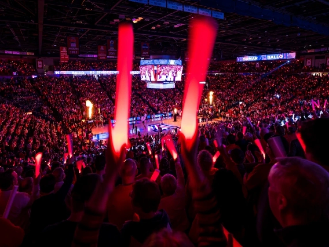 A crowd of people attend of basketball game in an indoor arena. The crowd is holding red, illuminated plastic sticks.