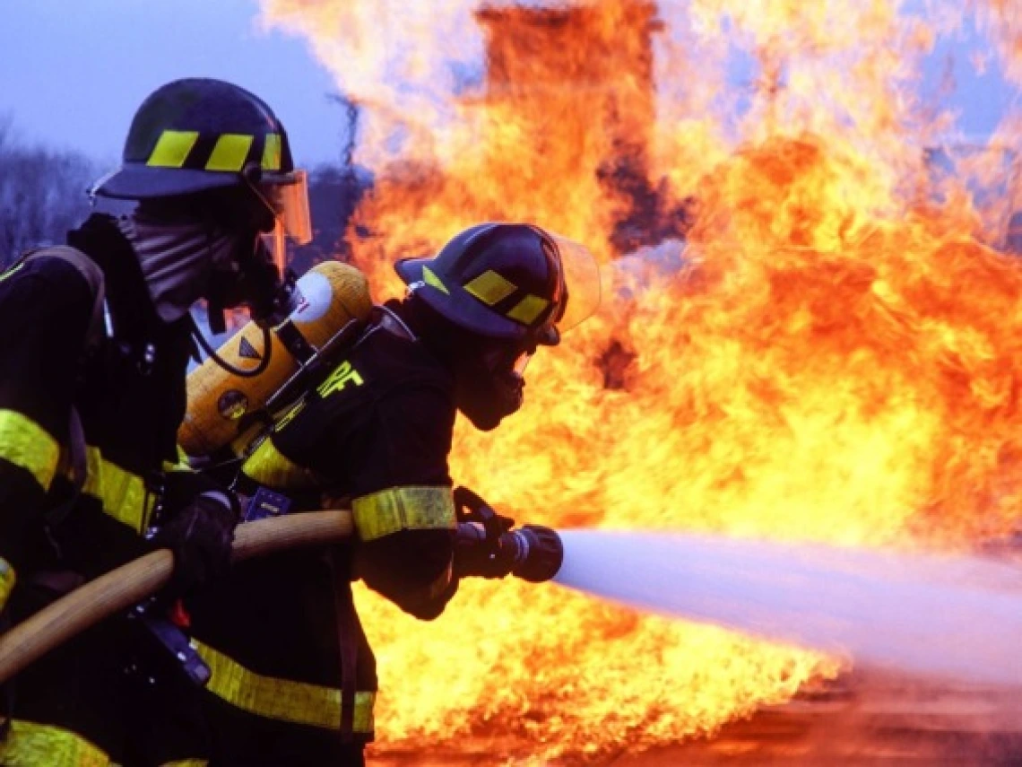 A pair of firefighters in full protective equipment hold a hose that is spraying water onto an active fire