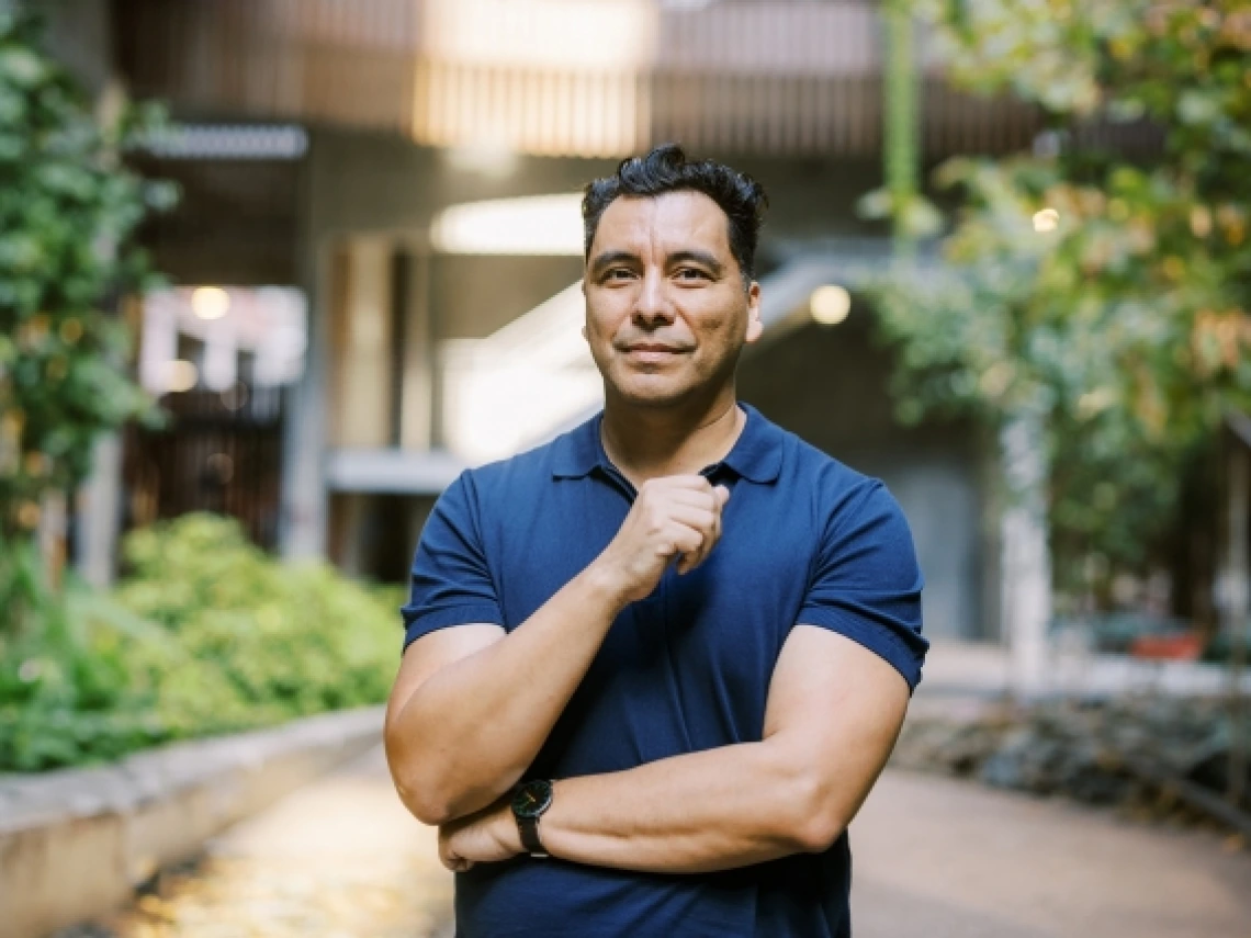 A man wearing a blue shirt stands with his arms crossed and one hand below his chin.