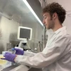 A young male scientist with curly brown hair, wearing a white lab coat and purple gloves, works inside a biosafety cabinet in a laboratory. He is handling a clear plastic container while surrounded by lab equipment, including a microscope and various pipette tip boxes. A reflection of his face is visible on the cabinet's glass, and a few coat hangers are sketched on the whiteboard behind him.