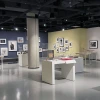 A modern gallery space at the Center for Creative Photography featuring an exhibition of framed photographs and archival materials. The walls are painted in dark blue, soft yellow, and white, with artworks arranged in salon-style groupings. Display cases throughout the room contain books, prints, and other photographic items under glass.
