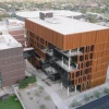 An aerial view of a copper-sided building with multiple levels of outdoor balconies. The building is attached to another via a walkway. Cars can be seen driving on the street outside.