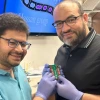 Two smiling researchers in a lab, both wearing glasses and gloves, hold up a small experimental circuit board labeled 'Graphene.' Behind them, a screen displays the words 'Hassan group' along with colorful molecular imagery. The setting suggests a breakthrough in advanced electronics or transistor technology.